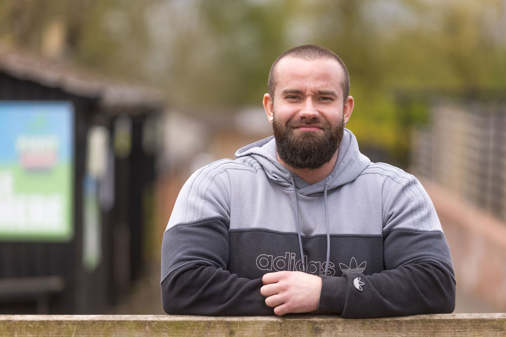 Former Public Services student, now Public Services teacher Marek Piontkowski leaning against a wooden gate wearing a grey hoodie.