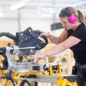 Female student using a buzzsaw, wearing ear protectors.