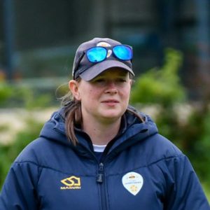 Female cricket coach, Emma Thatcher, wearing a blue puffer jacket and a baseball cap.