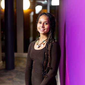 Young female, Samantha Rosser, wearing a black top stood against a purple wall.
